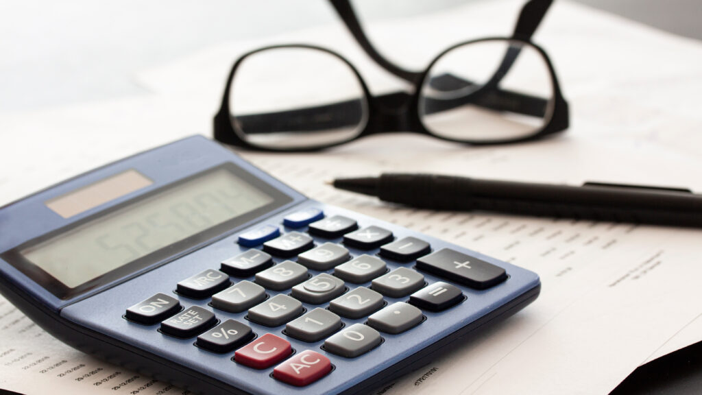 A calculator sits on some financial documents with a pen and eye glasses next to it.