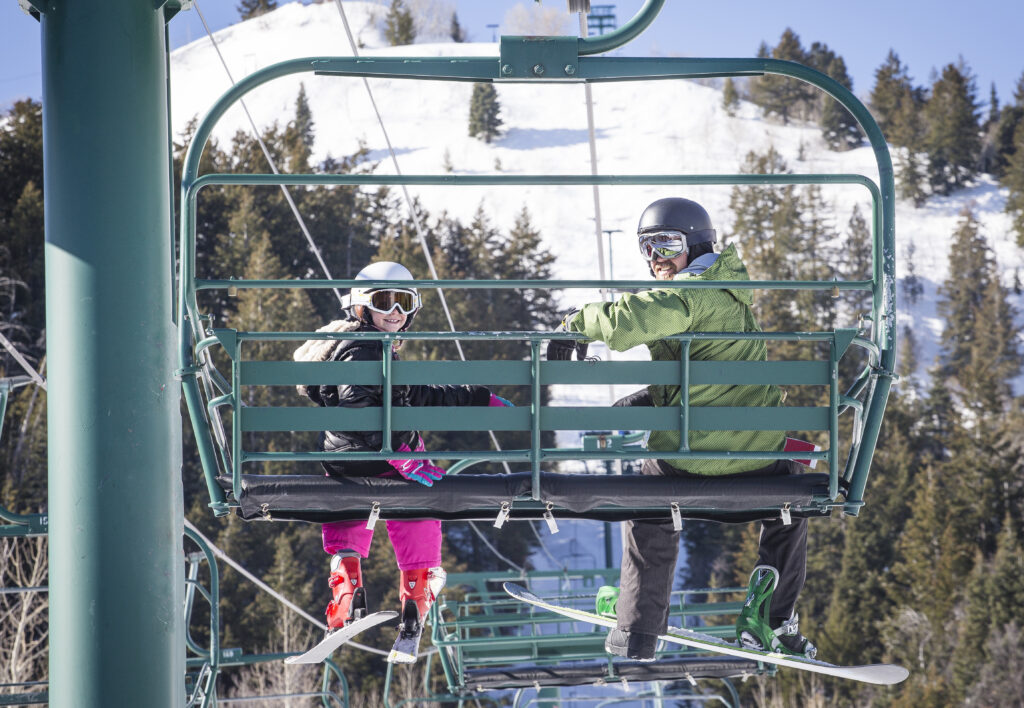 Smiling father and daughter riding a chair lift together on a sunny day at a ski resort.